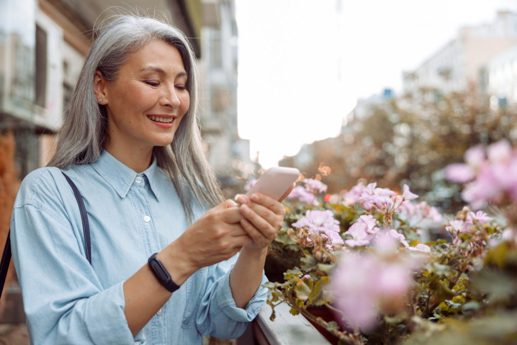 Smiling grey haired Asian woman uses cellphone standing on outdoors terrace